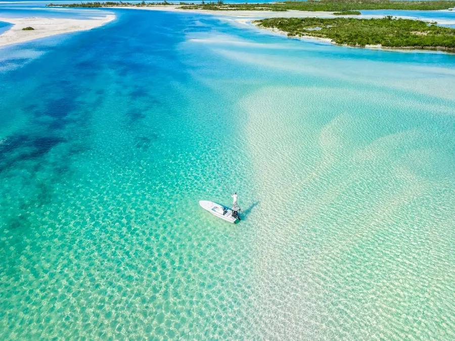A flats skiff poling across the clear turquoise bonefish flats of Acklins from the air, mangrove cays behind