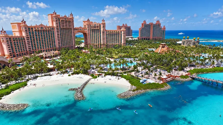 The pink resort towers on Paradise Island from the air, the archway between them, palm shaded pools and a white sand beach on turquoise water with paddleboarders offshore