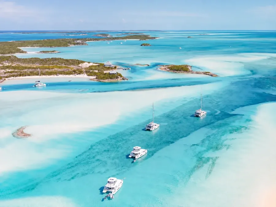 Sandbars drying out of turquoise shallows in the Bahamas, seen from the air, with four catamarans anchored along the deeper channel between them