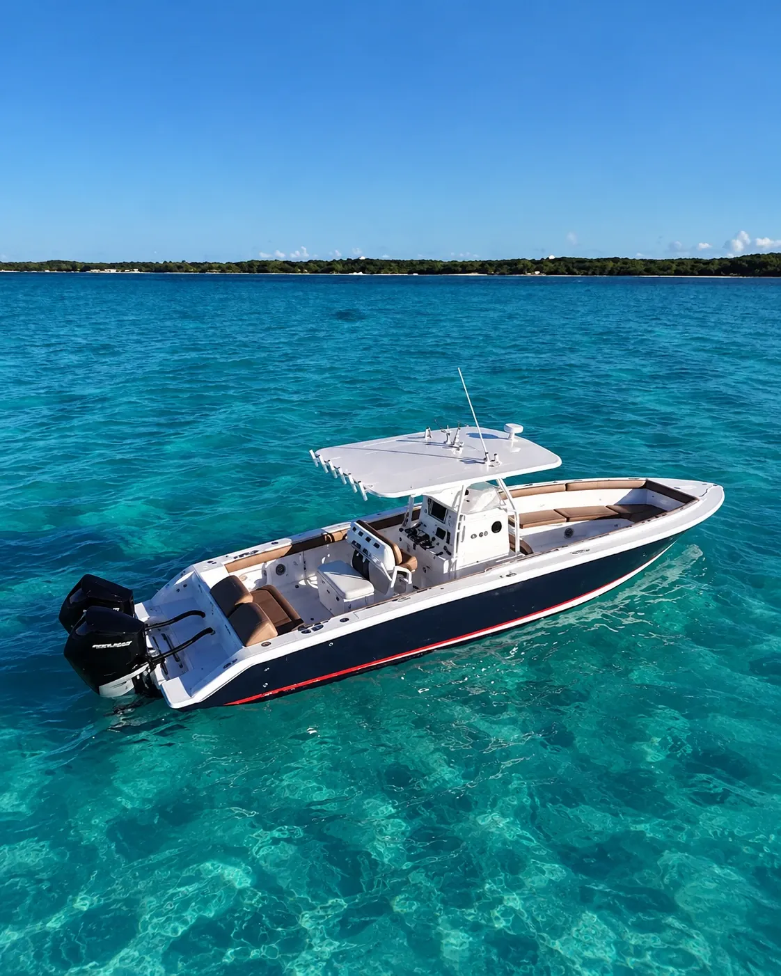 A navy center console boat with twin outboards anchored over clear turquoise water off a green island shore