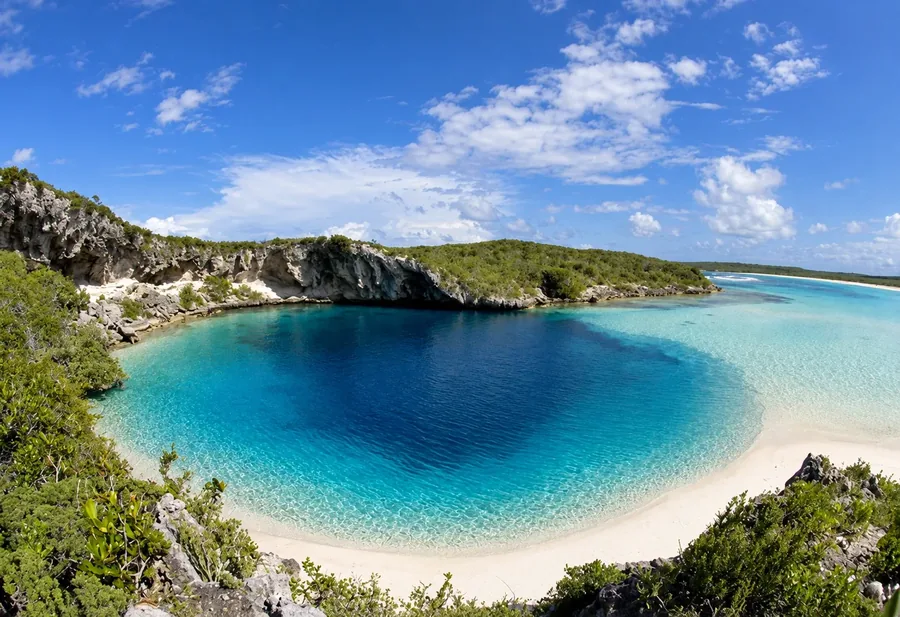 Dean's Blue Hole on Long Island seen from the cliff above, a deep circle of dark blue ringed by a crescent of white sand and turquoise shallows