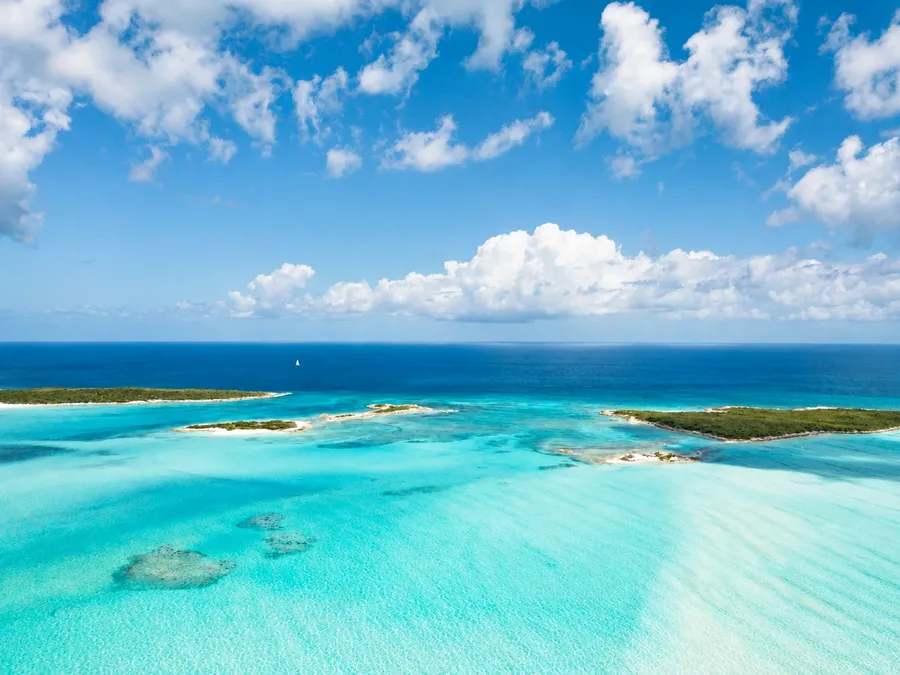 Small green cays and turquoise shallows at Great Guana Cay from the air, deep blue ocean and a sailboat on the horizon