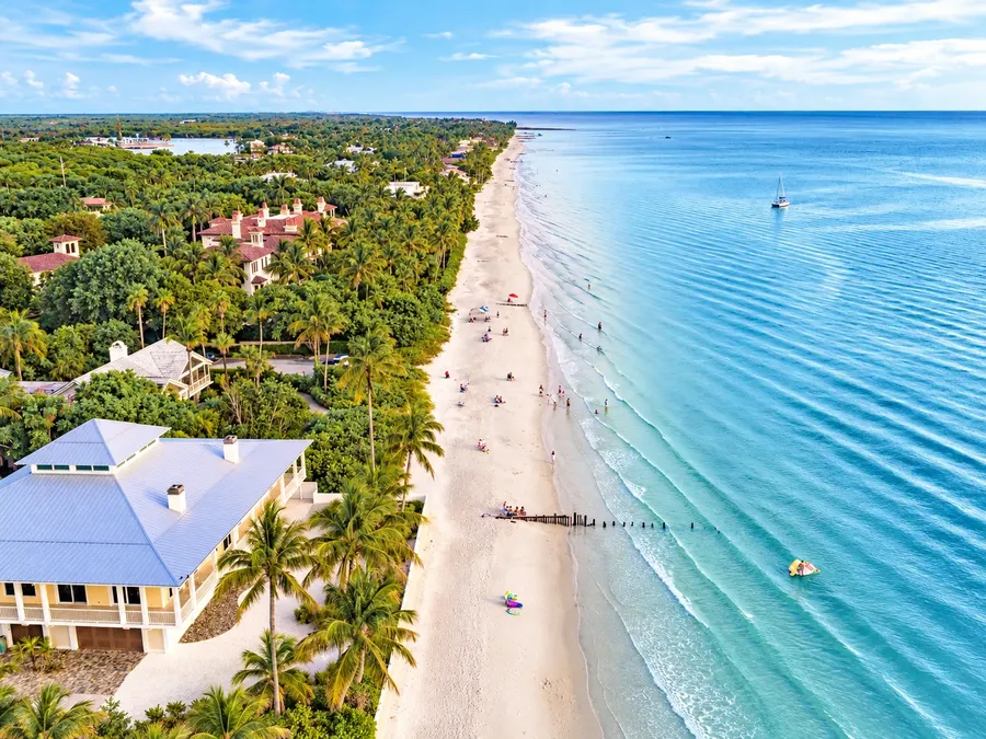 A long white beach on Florida's Gulf Coast from the air, palms and homes along the shore and calm blue water beside it