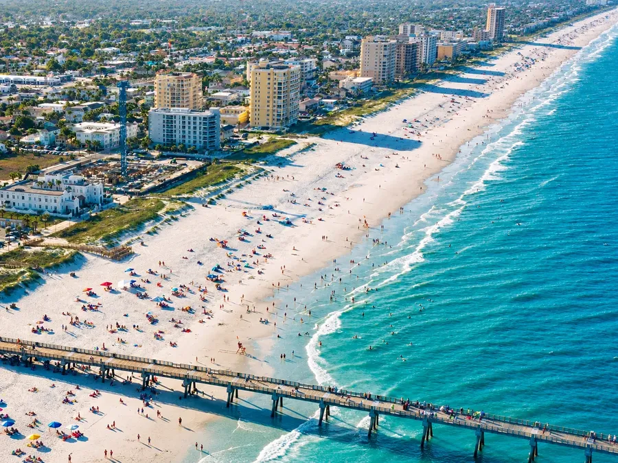 Jacksonville Beach and its pier from the air, a wide white beach with hotels along the shore and blue Atlantic surf