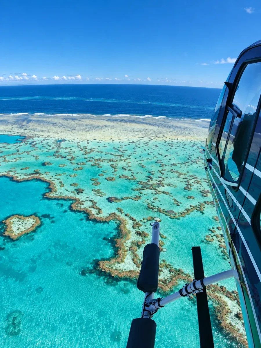 The view past the open door of a helicopter over a shallow reef: coral heads and sandbars in turquoise water, the pale reef edge and the open ocean beyond
