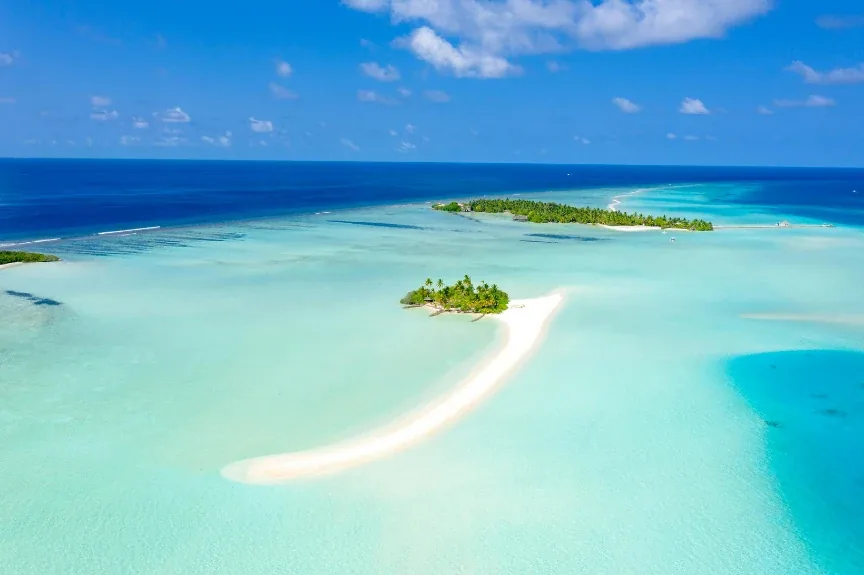 A pale sandbar curving away from a small palm island in turquoise shallows, the open ocean behind it