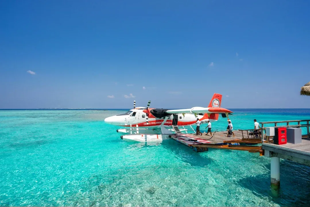 A seaplane on floats moored alongside a wooden dock in clear turquoise shallows, guests and ground crew walking out to meet it