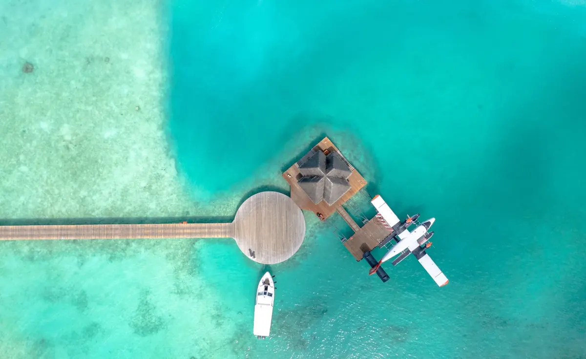 A seaplane on floats tied up at a wooden dock beside a boat, seen from straight above in clear turquoise water