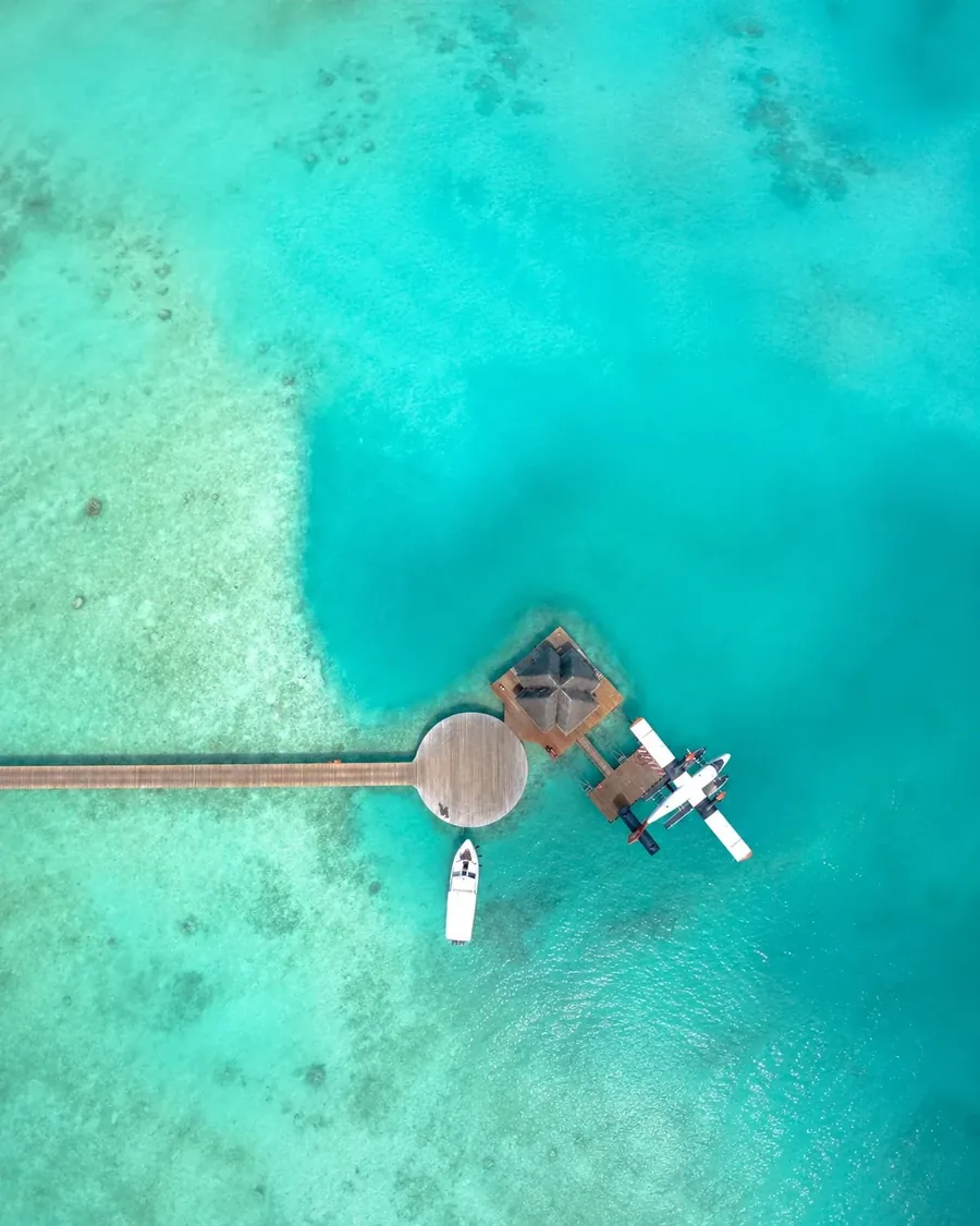 Seen from directly above, a seaplane on floats at the end of a jetty over turquoise shallows, a small boat moored alongside
