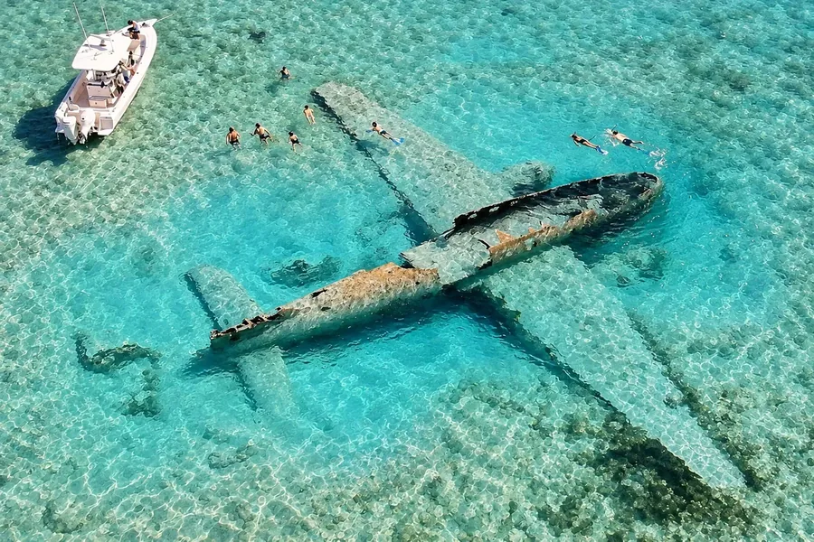 The sunken aircraft off Norman's Cay from the air, wings and fuselage lying in clear turquoise shallows with snorkellers and a boat beside it