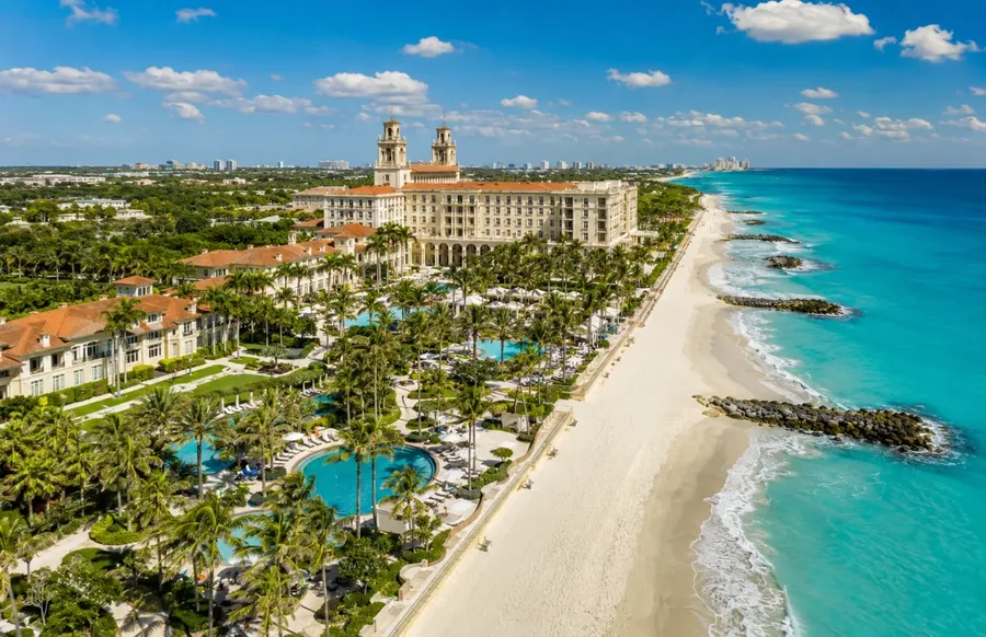 The Palm Beach oceanfront from the air, a large resort with red tile roofs and palm gardens above the white sand